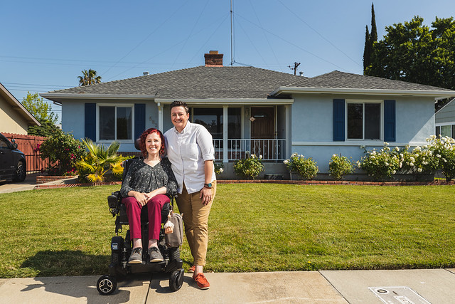Photo of red haired white woman in a wheelchair wearing a floral top and burgundy pants with a short haired white woman wearing an oxford shirt and khaki pants standing next to her with her arm around her. They are standing in front of a 1950s style one story blue house.