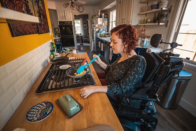 White woman in a wheelchair cooking eggs on the stovetop that she is able to roll under.