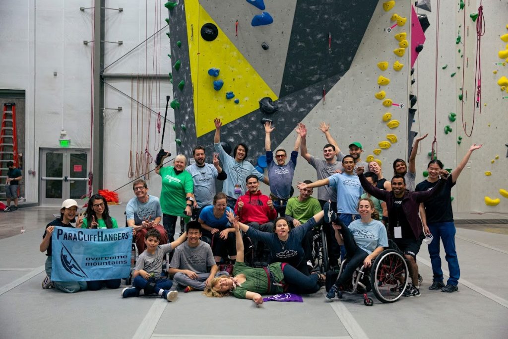 A diverse group of people, some in wheelchairs and some with wrist crutches pose in front of a colorful indoor rock wall. a couple of people hold up a cloth banner reading "ParaCliffHangers - Overcome Mountains"