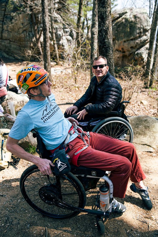 Two people in wheelchairs are looking comfortable and hanging out in an outdoor setting with large boulders and trees in the background.