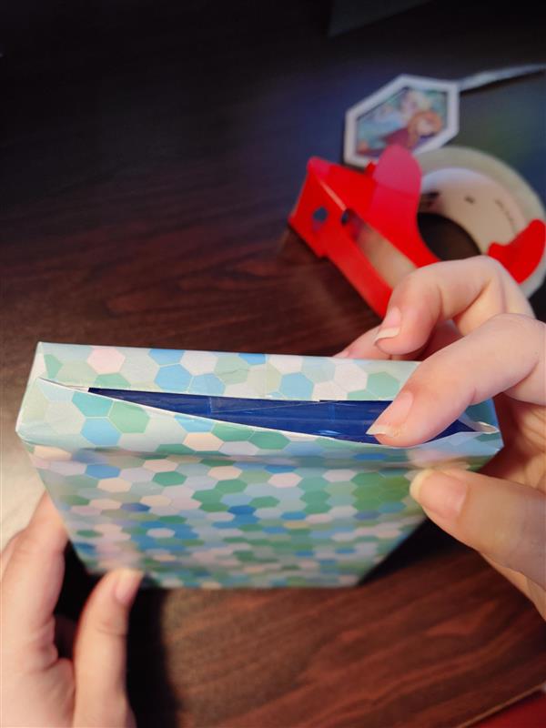 Hands hold a wrapped gift with a finger holding open the top untaped seam. There is a frozen themed gift tag and a roll of packing tape in the background.