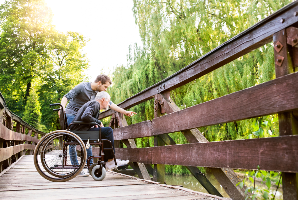 A person in a wheelchair and a person standing beside them are in the middle of a wooden bridge, surrounded by trees, look over the edge of the bridge to see the water down below.