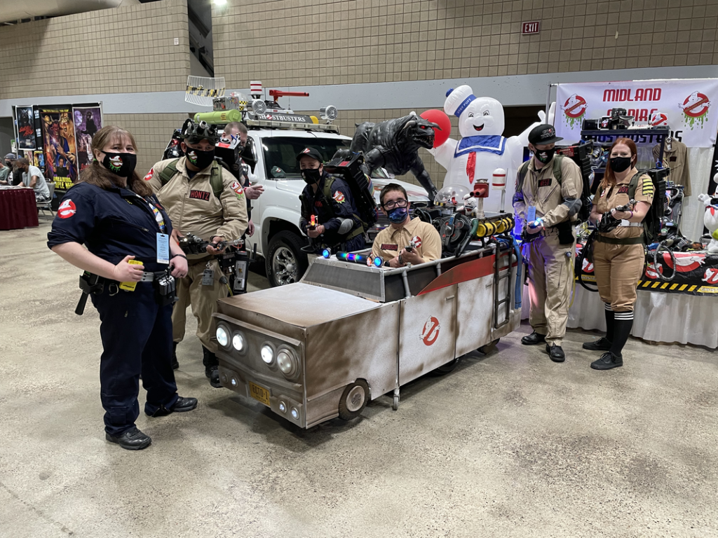 Someone seated in a Wheelchair posing for a photo. The wheelchair decorated as Ghostbuster car, around them are a group of people dressed up in Ghostbuster cosplay costumes.  