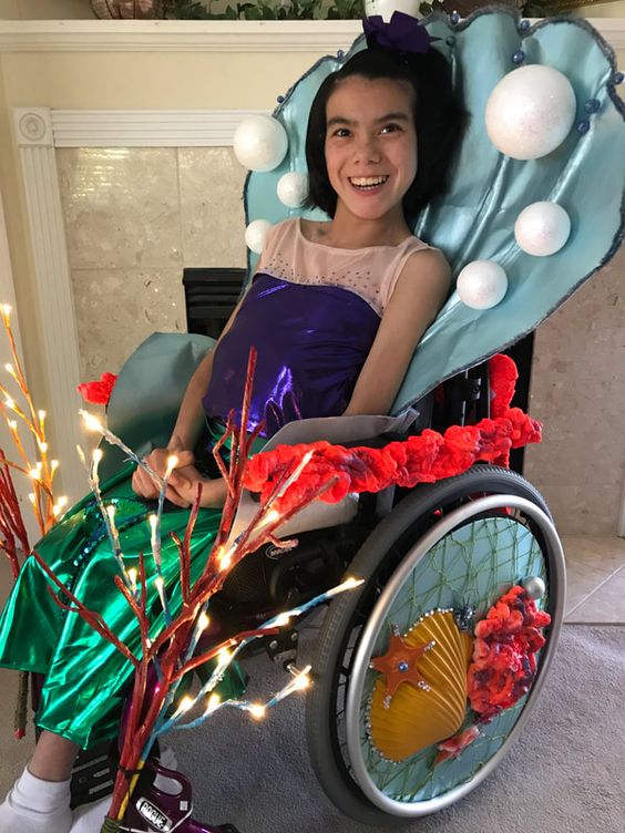Smiling girl poses for photo she is dressed in a mermaid costume is seated in a wheelchair. The back rest and wheels are decorated in sea shells and coral.  