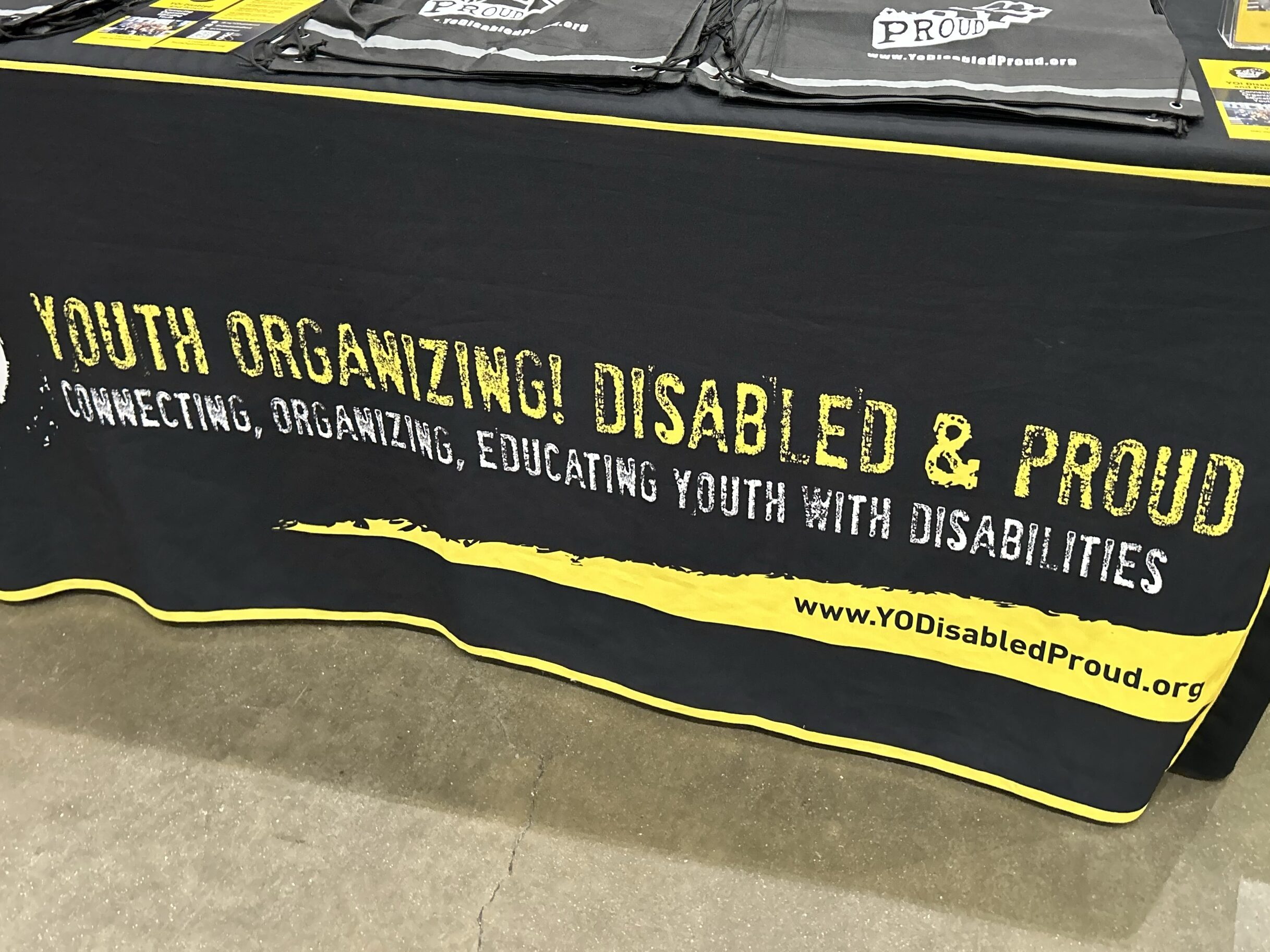 A table with a black and yellow table cloth displays black bags yellow flyers. The words "Youth Organizing! Disabled & Proud Connecting, Organizing Educating Youth with Disabilities
www.YODisabledProud.org"
are across the front of the table cloth.