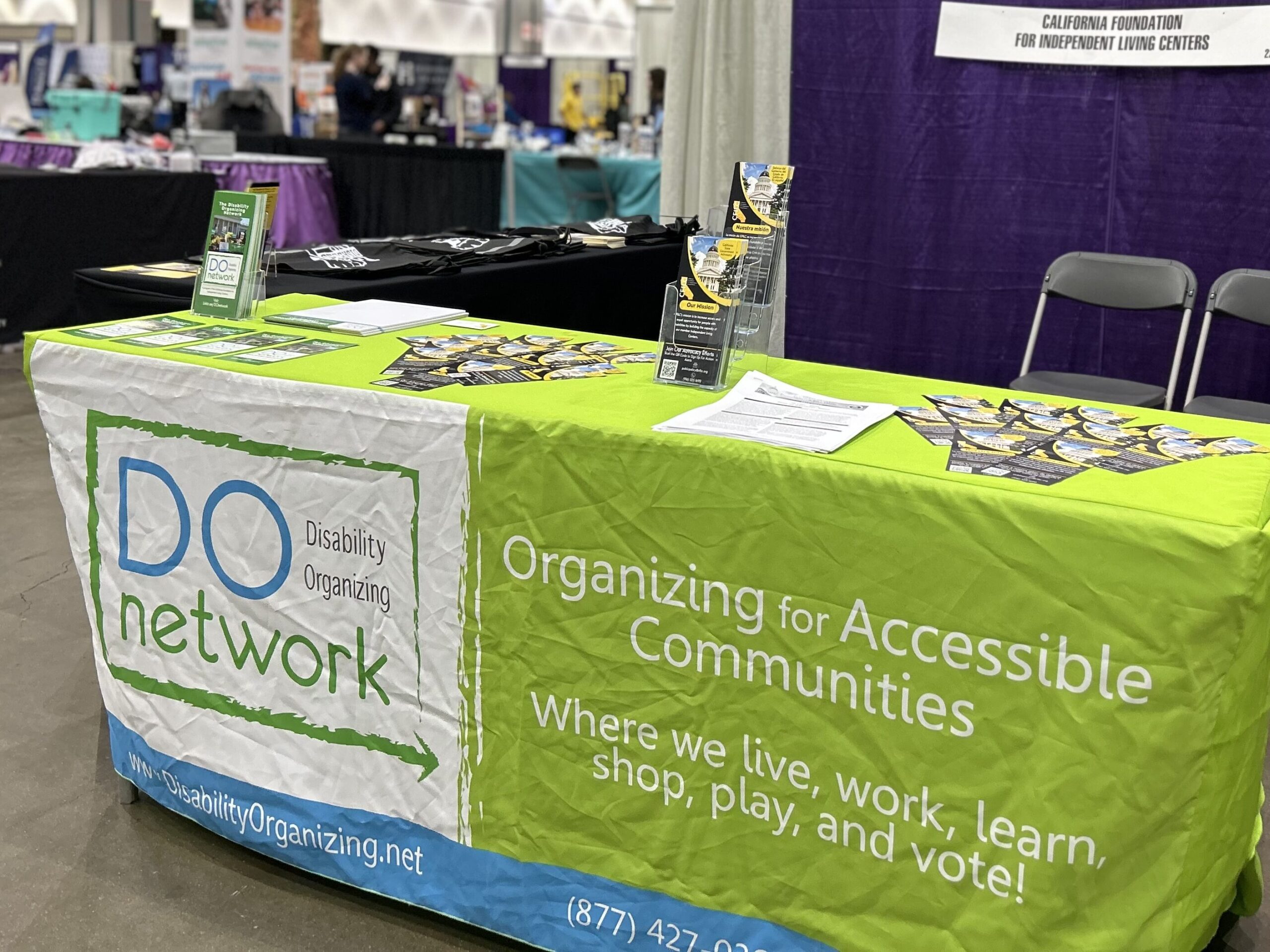 A table with a bright green, blue and white tablecloth displays flyers and sheets of paper. The words "DO Network Disability Organizing) and on the green is "Organizing for Accessible Communities Where we live, work, learn, shop, play and vote!" are across the front of the tablecloth.