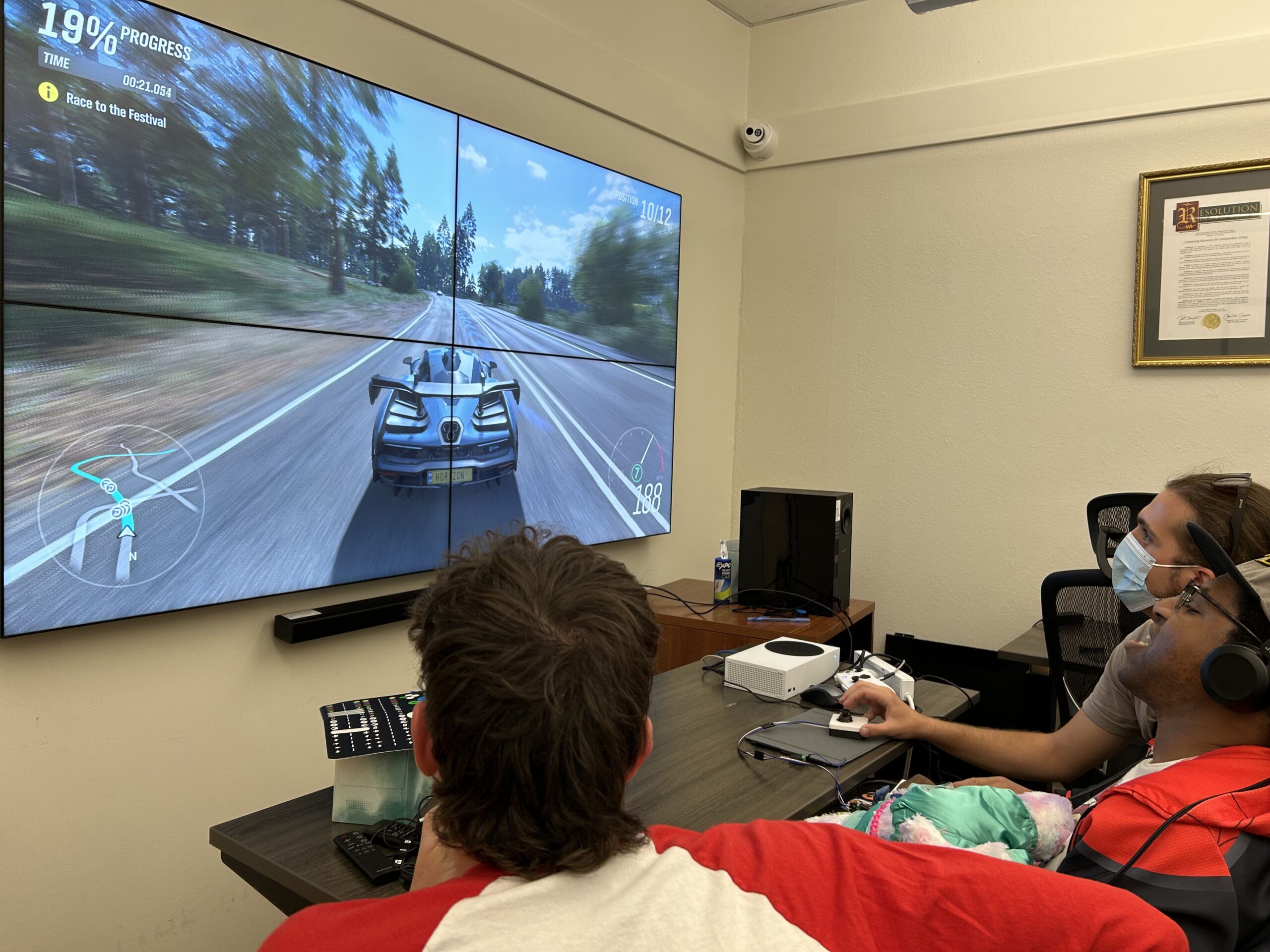 Three men play a racing game on a large screen. One is handling a controller. On the table in front of them are controllers to a video game unit.